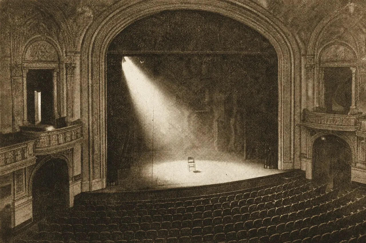 The interior of a grand old theatre, a single spotlit empty stage seen past rows of seats.